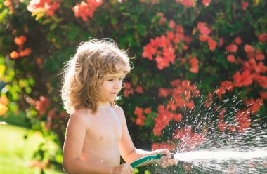 Çocuk bahçede oynuyor, hortumdan dökülüyor, yağmur yağıyor. Mutlu çocukluk kavramı. Çocuk ağaçlara su döküyor. Arka bahçedeki bitkilerle ilgilenen çocuk.