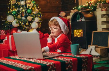 Noel hediyelerini online satın alın. Noel alışveriş konsepti. Hediye servisi. Küçük bir dahi. Noel Baba küçük yardımcı. Noel ağacının yanında dizüstü bilgisayarlı çocuk. Küçük çocuk santa şapka ve kostüm eğleniyor.