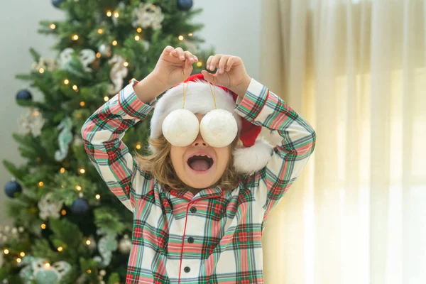Funny excited child with Christmas balls on eyas near Christmas tree at home. Child preparing for the Christmas and New Year. Child with bauble. Kid near Christmas tree. Decorated for winter holidays