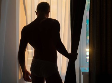 Seksi gey silueti. Adam soyunuyor. Çıplak gövdesi olan genç bir adam. Pencerede seksi, genç, kaslı bir adam var.