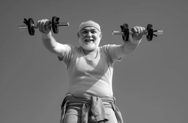 Senior man lifting weights. Senior man in gym working out with weights. Happy senior man with dumbbell looking at camera. Grandfather sportsman portrait on blue sky backgrounds