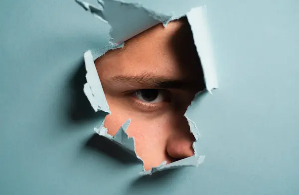 Man looking through paper. A jealous husband. Hole torn in paper with the eye of boy. Portrait of man looking through the hole in white paper. Distrustful look. Spy eye. Mans curiosity and gossip