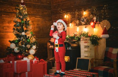 Mutlu küçük çocuk Noel Baba kıyafetleri giyiyor, Noel hediyesi kutusu da var. Noel konsepti. Şömine arka planı. Tatiller