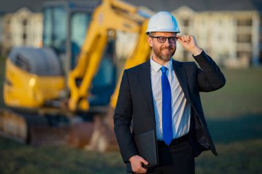 İnşaat mühendisi inşaat alanında çalışıyor. Olgun mühendis. İnşaat alanında takım elbiseli ve kasklı bir adam var. Orta yaşlı inşaat mühendisi kazıcının yanında duruyor.