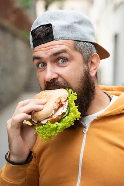 ひげ付きの男はファーストフードを食べる バーガーは役に立つ食べ物ではない とてもお腹がすいた男 ハンガーマンは屋外でハンバーガーを食べる 通りで美味しいハンバーガーを食べる ランチでバーガー チーズバーガーやハンバーガー — ストック写真