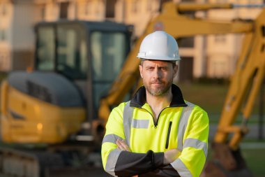 İnşaat alanında inşaat için kazı yapan bir inşaat işçisi. Binaların arka planında inşaat işçisi olan bir makine traktörü. Kazıcı traktör ve inşaat işçisi. İnşaat alanında miğferli bir inşaatçı