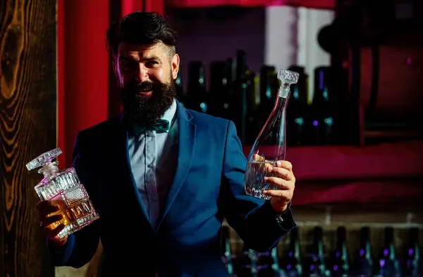 Handsome bearded barman with long beard and mustache with serious face made alcoholic cocktail in vintage suit on bar background