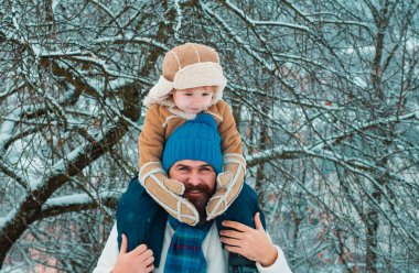 Baba oğlunu parka geri götürüyor. Çocuk babasının omuzlarında oturur. Baba ve oğul kış parkında eğleniyor.