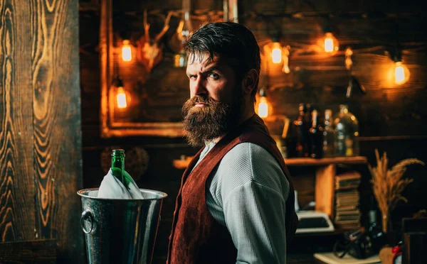 Handsome bearded barman with long beard and mustache has stylish hair on serious face. Man in vintage waistcoat. Brutal waiter. Vintage interior. Swag waiter with beard holds bucket with champagne in