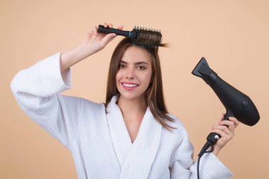 Woman in bathrobe combing hair, drying hair with hairdryer. Portrait of female model with a comb brushing hair. Girl with hairs brush and blow dryer. Hair care and beauty. Morning routine