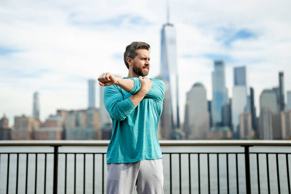 Senior man in sportswear stretching his arms outside in NYC background. Active senior man is exercising outdoor. Active after retirement. Mature retired sportsman doing stretching exercises