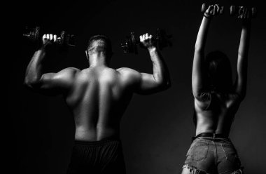 Çift halterlerle birlikte egzersiz yapıyor. Çıplak vücutlu kaslı bir adam, arka planda koyu renkli halterleri olan fitness kadını. Seksi, formda bir vücut. Dambılı çift eğitimi