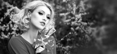 Bahar kadını pankarta karşı. Kıvırcık sarı saçlı, mükemmel tenli ve pembe çiçekli bir yaz bahçesinde güzel makyajlı güzel bir kadının yakın plan portresi.