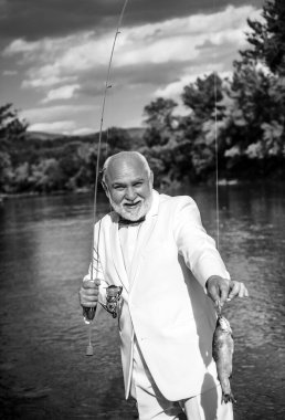 Portrait of cheerful senior man fishing. Grandfather with catch fish. Mature man fisherman in white suit and bowtie with fishing rod, spinning reel on river. Catching trout fish