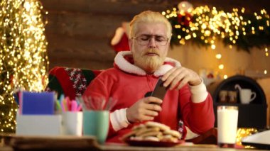 Evde Noel Baba kostümü giymiş bir baba Noel ağacının yanında uzaktan kumandayla televizyon izliyor. Komik Noel Baba