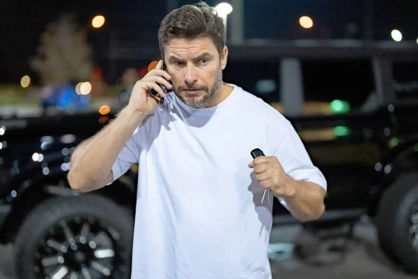 一个严肃的人在户外打电话 一个严肃的嬉皮士在夜市大街的汽车附近打电话 一个严肃的人在讲电话 严重英俊的嬉皮士看起来很野蛮 在电话里说话 — 图库照片