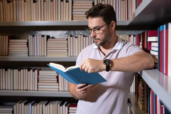 Portrait of teacher with book in library classroom. Handsome teacher in university library. Teachers Day. Teacher giving classes. School teacher in library. Tutor at college library on bookshelf