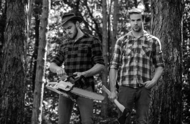 Woodcutters with chainsaw lumberjacks with axe. Hipsters men on serious face with axe. Lumberjack brutal and bearded holds axe. Two lumberjacks in forest.