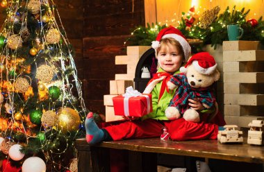 Oyuncak ayısıyla ve Noel hediyesiyle küçük mutlu çocuk kameraya bakıyor. Noel Baba şapkalı mutlu çocuk Noel 'i kutlar. Mutlu çocuk hediyeyle eğleniyor.