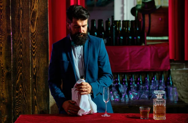Handsome bearded barman with long beard and mustache with serious face made alcoholic cocktail in vintage suede leather on purple red bar background