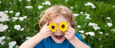 Bahar çocuğu suratlı pankart. Komik Çocuk ve Papatyalar. Çiçekli, mutlu, sarı saçlı çocuk. Gözleri papatya çiçeğiyle çimlerin üzerinde uzanıyor. Papatya gözlü çocuk, yazın yeşil çimlerin üzerinde.