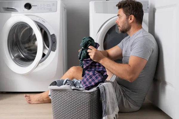 Man with clothes near washing machine. Laundry cleaning. Housework, homework, male housekeeper. Husband man doing laundry at home. Washer and dryer at home