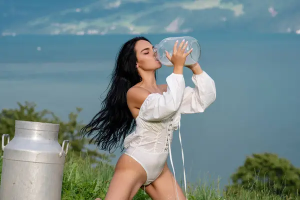 Sexy woman in lingerie drink milk from can and bottle against countryside. Sexy woman drinking milk in Alps. Sensual woman eat milk near Swiss Alps. Beautiful woman enjoys milk on alpine village