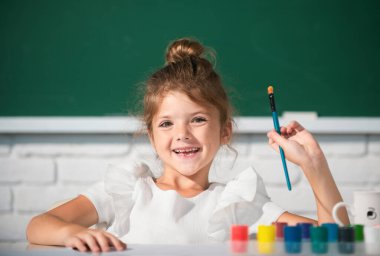 Child girl draws in classroom sitting at a table, having fun at school blackboard background. Painting school lesson, drawing art