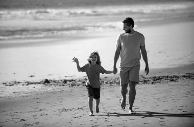 Baba ve çocuk el ele tutuşup birlikte yürüyorlar. Baba oğul yaz plajında yürüyorlar.