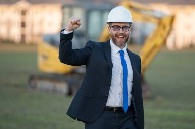 İnşaat işçisi yatırımcı. İnşaat alanının önündeki yatırımcı. Modern ev inşaatında başarılı yakışıklı bir adam. Takım elbiseli ve kasklı orta yaşlı yatırımcının portresi