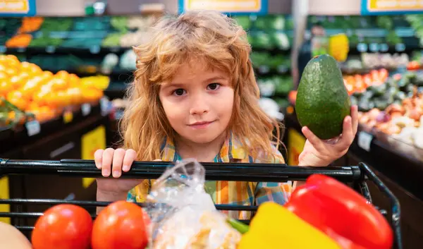 Child boy is shopping in a supermarket. Healthy food for young family with kids. Portrait of smiling little child with shopping cart full of fresh vegetables. Kids at grocery store or supermarket