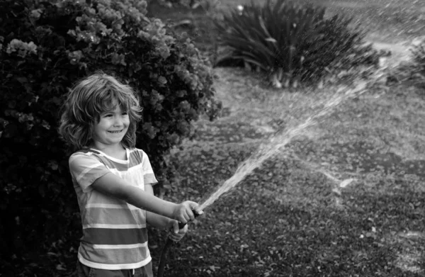 Happy kid boy pours water from a hose. American kids childhood. Child watering flowers in garden. Home gardening