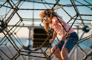 Kid boy climbing the net. Child playing on outdoor playground. Kids play on school or kindergarten yard. Kids play and climb outdoors on sunny summer day