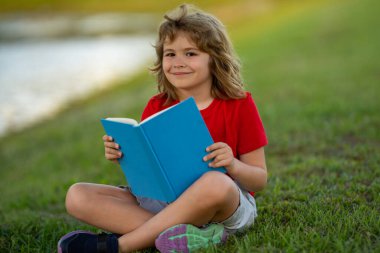 Yazlık parkta kitap okuyan sevimli bir çocuğun açık hava portresi. Sarışın çocuk çimlerin üzerinde oturup kitap okuyor. Park 'ta yaz tatili. Öğrenci çocuk kitapla çalışıyor, öğreniyor. Kitap deposu