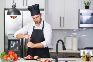 Önlüklü adam ve aşçı şapkalı adam mutfakta yemek pişiriyor. Mutfakta sağlıklı yemek pişiren yakışıklı adam. Adam mutfakta yemek pişiriyor. Taze yemek malzemeleriyle ev menüsü. Modern mutfak iç mimarisi