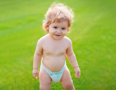 Yeşil çimlerin üzerinde, doğada oynayan, bez takmış küçük bir bebek. Çocuklar oynuyor. Bebek ve yaz güneşli hava. Küçük komik çocuk portresi. Sarışın çocuk, gülen duygu, gülen surat