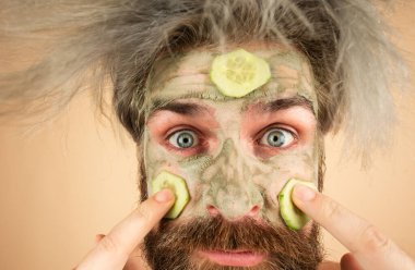 Yüzünde balçık maskesi ve salatalık dilimleri olan komik bir adam. Erkek spa, cilt bakımı, sağlık ve yüz bakımı konsepti. Adamın kozmetik nemlendirici maskesi var.
