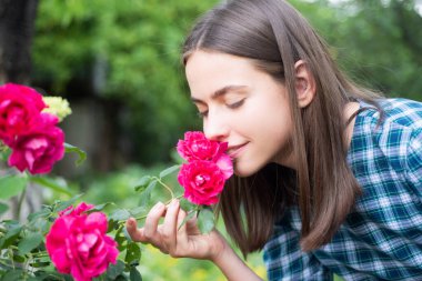 Beautiful girl smelling a rose flower in spring park. Young woman in flowering garden with roses. Beauty model with spring flowers. Happy spring. Pretty woman enjoying smell flowers in spring park