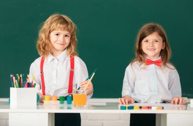 Okulun gülümseyen çocukları, kız ve oğlan boyaları, renkleri ve fırçaları okul karatahta arka planında, kameraya bakıyor.