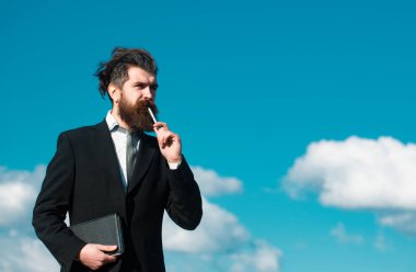 Gökyüzü olan bir iş adamı. İşadamı kalemle ilgili bir şey düşünüyor. İş hayalleri. Tişörtlü ve kravatlı düşünceli genç adam elinde not defteri ve kalemle