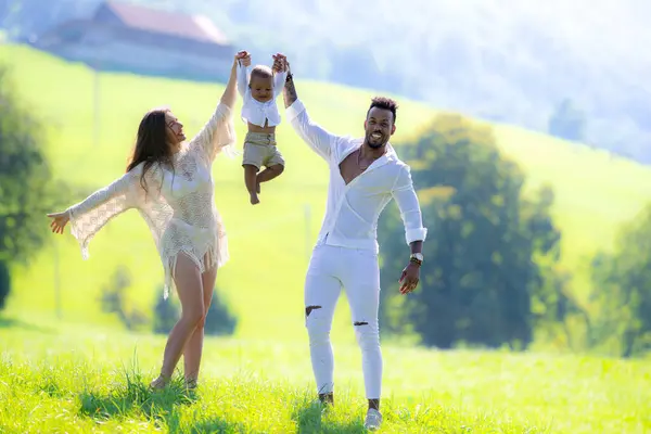 Young multi ethnic family portrait. Mixed race parents with baby spend time together hugs and kisses in park. African american husband and caucasian wife and baby interracial child enjoying summer day