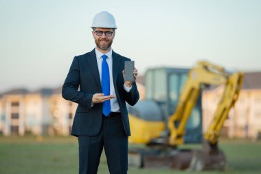 Başarılı bir inşaat işletmesi sahibi. Kazıcının yanındaki kask ve takım elbiseli inşaat işçisi. İnşaat alanının önünde kendine güvenen bir inşaat sahibi. İnşaat mühendisi.