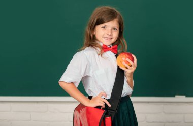 Çekici, küçük, neşeli bir kızın yakın plan portresi. İçerideki sınıftaki masanın üstünde oturuyor. Komik liseli kız suratı. Eğitim, öğrenim ve çocuklar