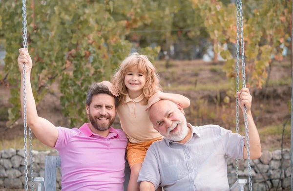 Three generations of men together, portrait of smiling son, father and grandfather swinging on the swing, having fun in the park outdoor