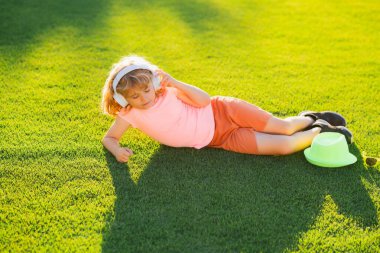 Child enjoy listens to music in headphones, laying on grass, dreaming and relaxing. Cute and happy child 7-8 years old lying on the grass and listening to music in headphones on a sunny summer day