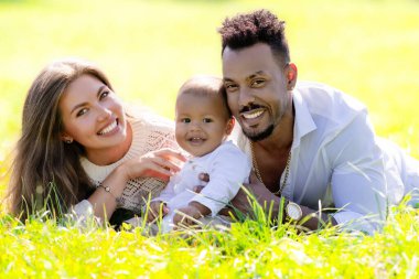 Çok ırklı ebeveynler ve parkta melez bir bebek. Çok ırklı aile portresi. Bahçede melez bir bebek var. Çeşitli ailelerden. Afrika kökenli Amerikalı baba beyaz anne ve bebeği çimenlerde yatıyor.