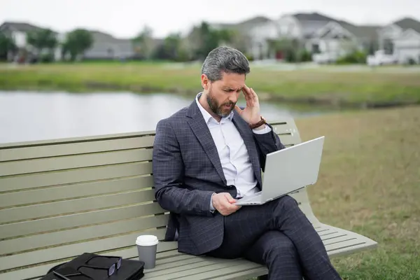 Stressed business man in suit sitting on bench. Stressed ceo manager work on laptop. Business man have stress and headache. Head pain migraine. Tired freelancer man online work from laptop in park