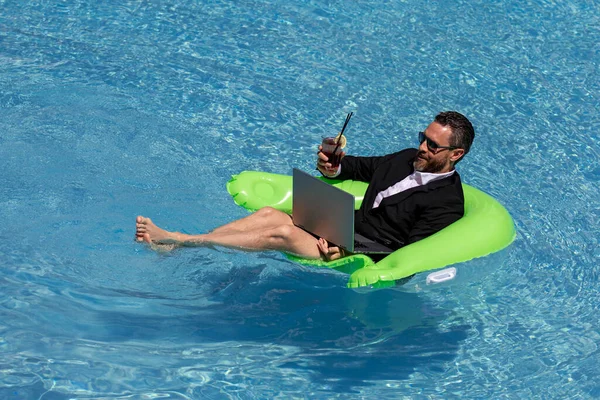 Funny business man in a business suit floating in the water in the pool. Remote summer work online. Crazy freelancer. Business and summer. Business man drink summer cocktail and using laptop in pool