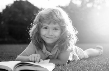 Çimlerin üzerinde uzanmış kitap okuyan çocuk. Günlük giysiler içinde, kitap okuyan ve parkta çimlerin üzerinde yatarken gülümseyen sevimli küçük bir çocuk.