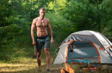 Yakışıklı ve kaslı oduncu elinde baltayla kamp yapıyor. Baltalı bir adam doğal manzaradaki şenlik ateşine bakar. Ormandaki ateşin yanındaki adam. Seyahat, kamp gezisi.